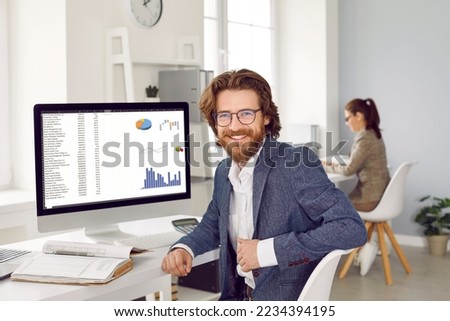 Happy male financial accountant or data analyst at work in office. Handsome bearded young man in suit and eyeglasses sitting at computer screen with graphs and charts, looking at camera and smiling