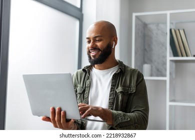 Happy Male Freelancer Using Laptop Computer And Wearing Earbuds, Standing Near Window And Typing On Keyboard At Home. Technology And Freelance Career Concept