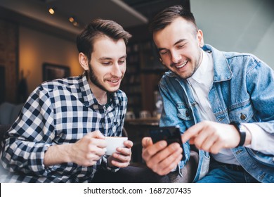 Happy Male Friends In Stylish Casual Clothing Watching Interesting Video On Smartphone And Holding Coffee Cup While Sitting In Cafe