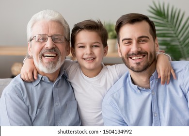 Happy male intergenerational 3 three generation men family close up portrait, cute child boy son grandson embracing young grown father and old grandfather laughing bonding looking at camera at home