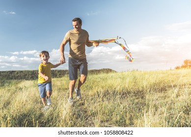 happy male with kite and boy running on sunlit meadow and holding hands. Full length