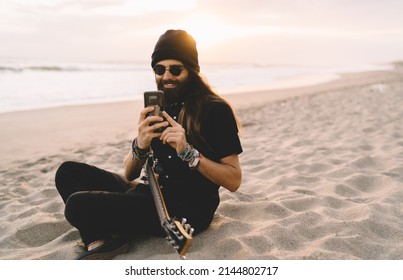 Happy male musician in sunglasses using cellphone technology for chatting and messaging at coastline beach, carefree hipster guy with six-string hawaiian guitar shooting video on smartphone gadget