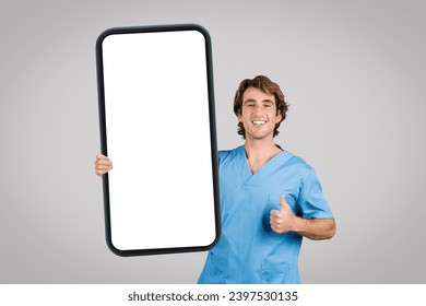 Happy male nurse in blue scrubs giving a thumbs-up sign while holding an oversized blank smartphone frame, ideal for app or telehealth promotion, on grey background - Powered by Shutterstock