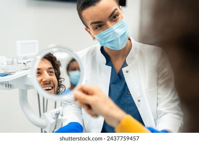 Happy male patient looking at mirror after dental treatment In a clinic. Cheerful man enjoying his beautiful smile. Female orthodontist communicating with satisfied patient after successful procedure. - Powered by Shutterstock