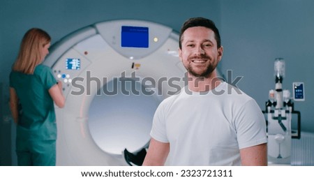 Happy male patient at well-equipped MRI laboratory, Portrait of cheerful man at modern tomography room. Man smiles with teeth at background of MRI capsule. Female doctor adjusts CT scanner behind man.