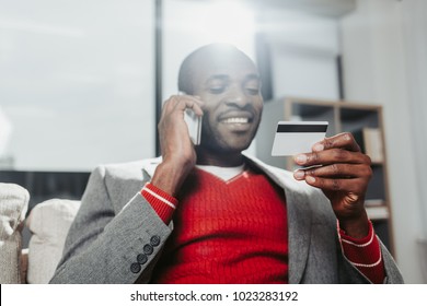 Happy male person relaxing on divan and talking by mobile phone. He is looking at payment card in his hand with joy Stock Photo