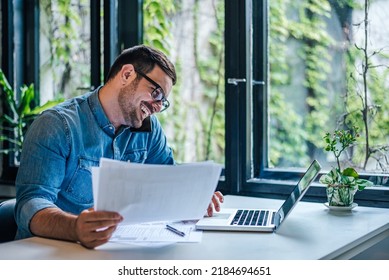 Happy Male Professional Discussing On Smart Phone While Using Laptop. Male Entrepreneur With Documents Is Multi Tasking At Table. He Is Wearing Casuals At Home.