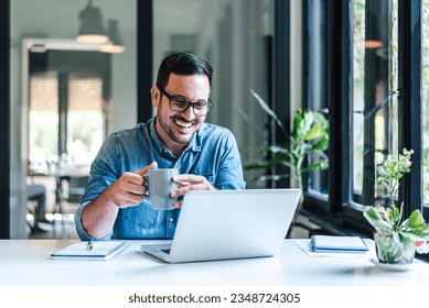 Happy male professional having coffee. Young smiling businessman looking at successful business plan on laptop. He is working at table by window in his home or office. - Powered by Shutterstock