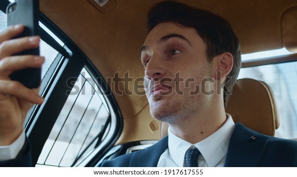 Happy male professional telling good news in video\
message on smartphone in business car. Cheerful business man making\
selfie video on phone in automobile. Smiling man talking to video\
chat in car.