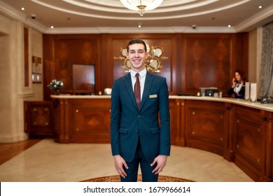 Happy male receptionist worker in uniform looking at camera with a smile while standing at hotel lobby. Horizontal shot