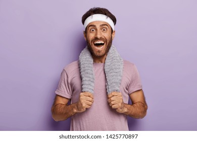 Happy Male Runner Smiles Broadly, Surprised To Cover Long Distance On Marathon, Has Towel On Neck, Wears Casual T Shirt And White Headband, Isolated Over Purple Background. Sport And Training Concept