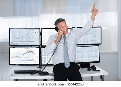 Happy Male Stock Trader Pointing Upwards While Using Telephone At Desk In Office