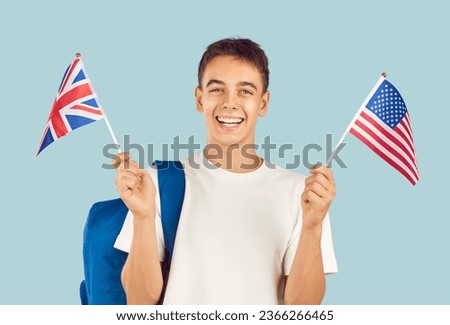 Happy male student learning English language. Studio portrait of school boy with backpack holding American and British flags, looking at camera and smiling isolated on light blue background