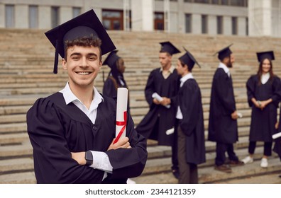 Happy male student on graduation day. Portrait of a young man in a black square cap and gown standing outside his school, college or university, holding his diploma, looking at the camera and smiling - Powered by Shutterstock