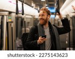 Happy male subway passenger is standing in the train wearing wireless headphones and a smartphone in his hand