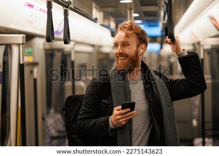 Happy male subway passenger is standing in the train wearing wireless headphones and a smartphone in his hand