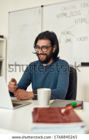 Happy male teacher sitting at his workplace and teaching online, looking at computer screen and smiling. High quality photo