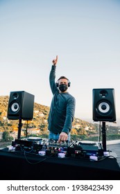 Happy Male Techno DJ, Wearing Black Protective Face Mask, With One Arm Raised, Playing Music In A Private Party At A Terrace House, With Nice Mountain Views. Outdoors, Sunset, Daytime.