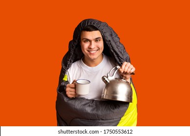 Happy Male Tourist Covered In Sleeping Bag Holding Tea Pot And Mug, Orange Studio Background