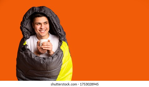 Happy Male Tourist Covered In Sleeping Bag Drinking Tea And Looking At Free Space, Orange Studio Background