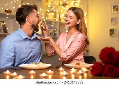 Happy man about to taste spaghetti playfully offered by woman's hand, both sharing a joyful moment during a candlelit dinner with roses. - Powered by Shutterstock