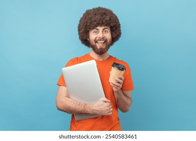 Happy man with Afro hairstyle standing with notebook and takeaway coffee in hands, finishing work, looking at camera with positive expression. Indoor studio shot isolated on blue background - Powered by Shutterstock