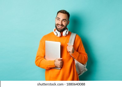 Happy man with backpack and headphones, holding laptop and smiling, looking left thoughtful, standing over turquoise background