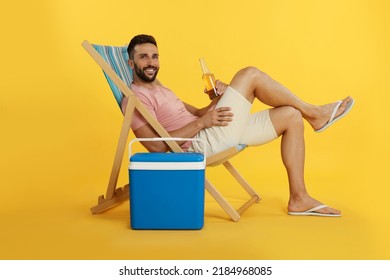 Happy man with bottle of beer resting in deck chair near cool box on yellow background