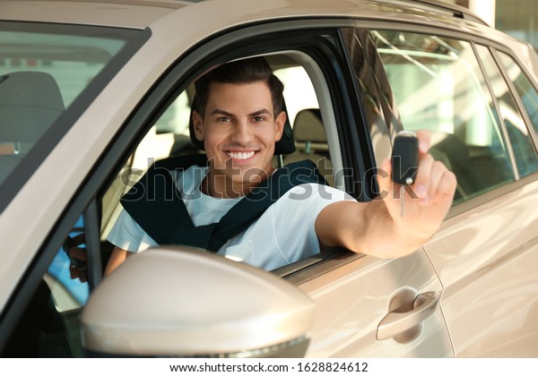 Happy man with car key sitting in modern auto\
at dealership