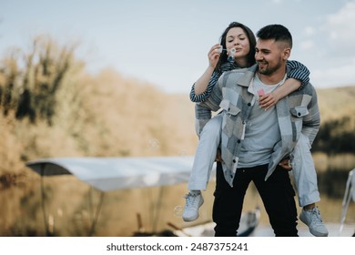 Happy man carrying woman on his back, blowing bubbles together in a natural lakeside setting, expressing joy and togetherness. - Powered by Shutterstock