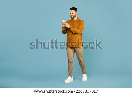 Happy man in casual wear using mobile phone chatting online, walking on blue background, full length, free space