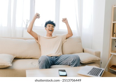 happy man celebrating success at home, sitting on a beige couch with raised arms, bright natural light, casual clothing, joyful expression, minimalistic interior design, excitement - Powered by Shutterstock