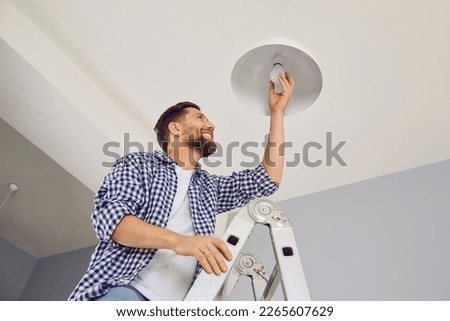 Happy man changing a light bulb at home. High angle shot of a young man in a checkered shirt standing on a step ladder and placing a modern energy-saving lightbulb in a white lamp on the ceiling