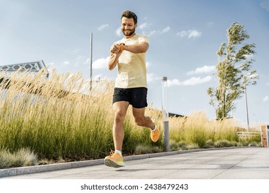 Happy man checking progress on fitness watch while running, tall grass background, sunny day.

