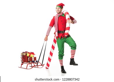 happy man in christmas elf costume carrying sleigh with presents and big candy cane isolated on white