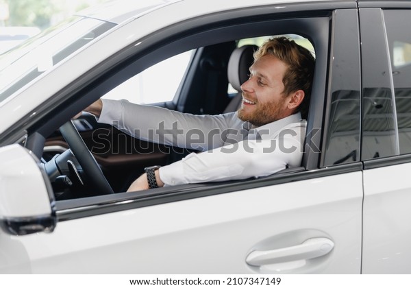 Happy man customer male buyer client in white\
shirt drive car hold wheel show thumb up choose auto want buy new\
automobile in showroom vehicle salon dealership store motor show\
indoor Sale concept.