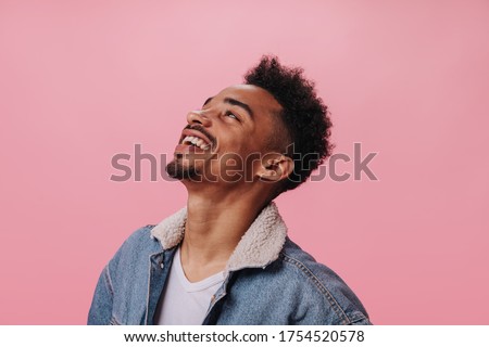 Happy man in denim outfit smiling on pink background. Cheerful brunette guy in white t-shirt laughing on isolated backdrop