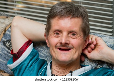 A Happy Man With Down Syndrome Leans Back On A Couch With A Big Smile.  He Is Older, And Has Lost All His Teeth.  He Is Looking At The Camera And Has Several Pens Attached To His Shirt.