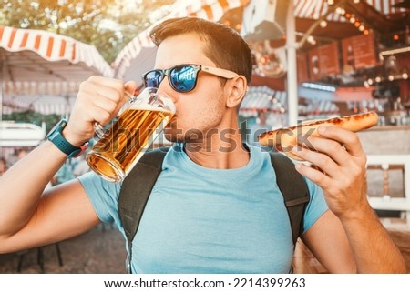 Happy man drinking beer and eating traditional german bratwurst - hotdog at funfair and street food festival. National cuisine and biergarten concept