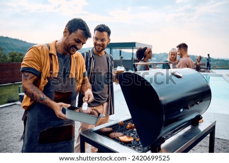 Happy man drinking beer and talking to his friend who is making barbecue at poolside party. 