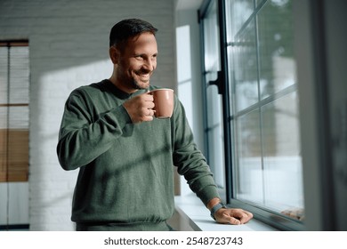 Happy man drinking tea while standing by the window at home and looking through it. Copy space.