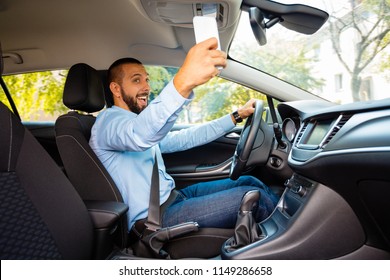 Happy Man Driving A Car And Taking Selfie With Mobile Phone At The Same Moment. 