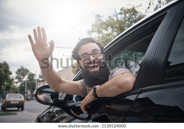 Happy man driving a car and\
waving through the opened window, saying hello. City positive\
people