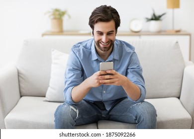 Happy Man In Earbuds Making Video Call On Smartphone Sitting On Sofa At Home. Guy Texting Using Phone In Living Room Indoor. Technology And Gadgets, Great Mobile Application Concept