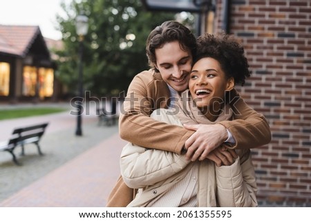 happy man embracing amazed african american girlfriend near building outdoors