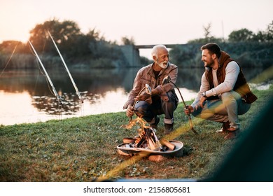 Happy man enjoying with his mature father while preparing fish on campfire during their camping day at sunset. - Powered by Shutterstock