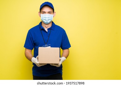 Happy Man With A Face Mask And Gloves Holding A Small Package Against A Yellow Background. Male Courier Making A Delivery During The Virus Pandemic 