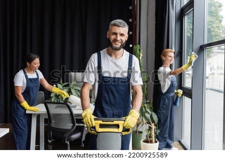 happy man with floor scrubber machine looking at camera near multiethnic women cleaning office