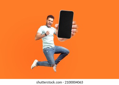 Happy man flying and jumping in air and showing big mobile empty screen for copy space and advertising area. indoor studio shot isolated on orange background - Shutterstock ID 2100004063