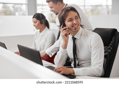 Happy Man In Formal Wear Adjusting Headset Microphone And Looking Away With Smile, While Sitting At Desk With Laptop Near Colleagues And Talking With Client During Work In Call Center Service